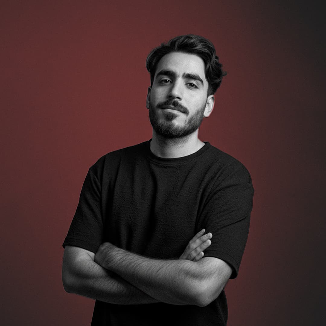 Homme barbu aux bras croisés portant un t-shirt noir devant un fond rouge dégradé.